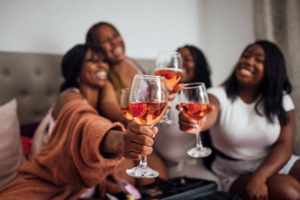 A group of female friends wearing pyjamas, spending the night together in one of their flats in Newcastle upon Tyne, England. They are all sitting on a bed, having a cheers with a glass of wine while smiling and the main focus is the wineglasses while the women are out of focus in the background.